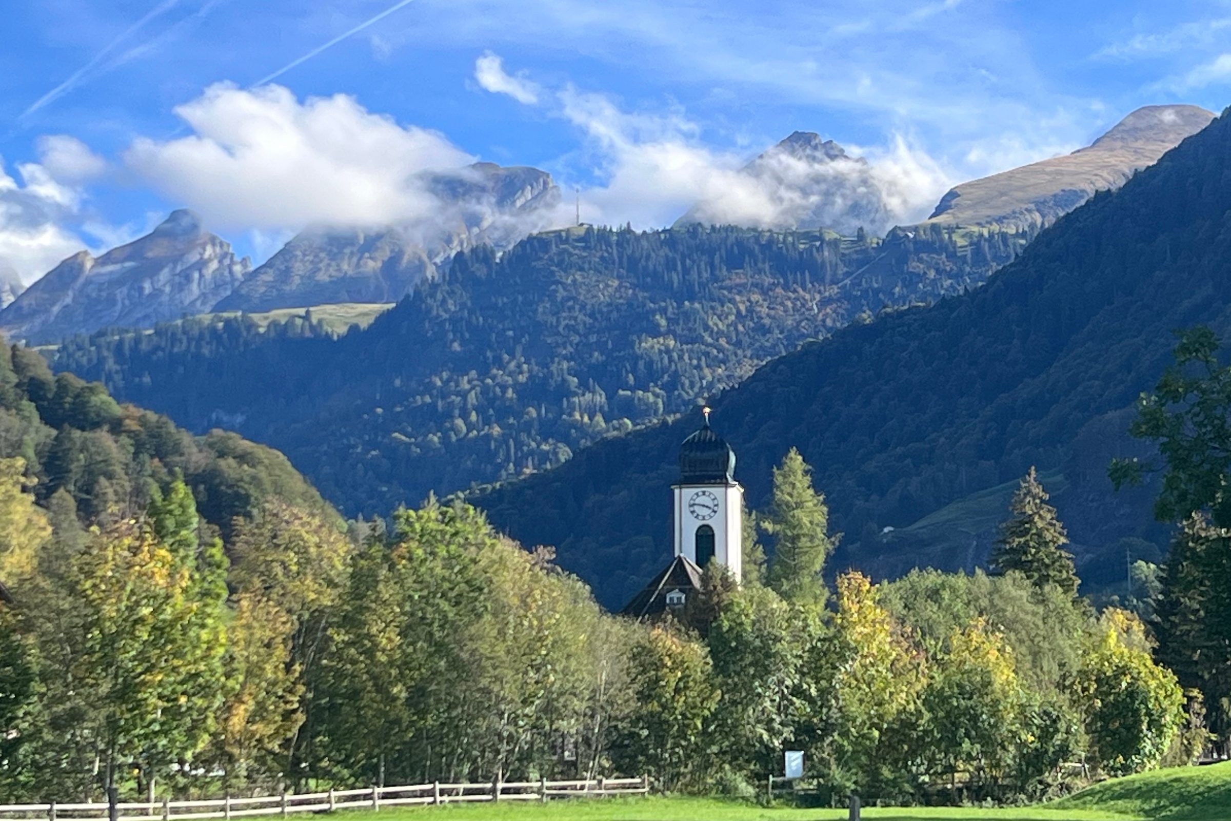Der Kirchturm von Neu St. Johann zwischen herbstlichen Bäumen, dahinter bewaldete Hänge und die Churfirsten in Wolken.